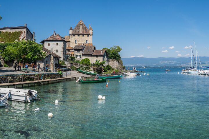Yvoire's pointed-roof castle overlooks stone houses, small fishing boats and a sailboat harbour on Lake Geneva beneath a clear sky.